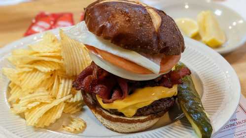 A hearty burger with bacon, cheddar, lettuce, and tomato, served with potato chips and a pickle on the side.