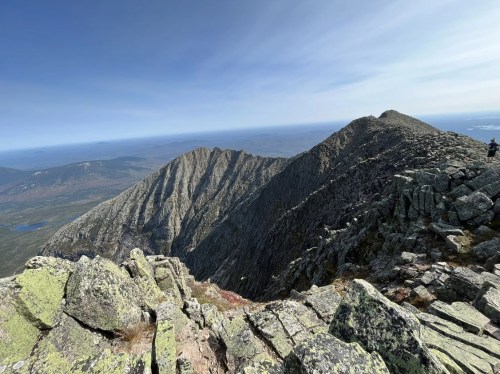 A panoramic view from a mountain summit, showcasing rugged peaks and a vast landscape under a clear blue sky.