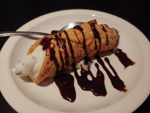 A cannoli filled with creamy filling, drizzled with chocolate sauce, served on a white plate with a spoon.