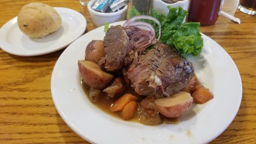 A plate of tender roast beef with potatoes, carrots, and lettuce, accompanied by a roll on the side.