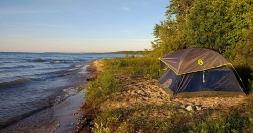 A tent set up on a sandy shore by a calm lake, surrounded by greenery and bathed in warm sunlight.