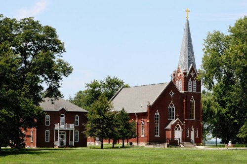 A red brick church with a tall steeple and cross, alongside a historic building, set in a green landscape.