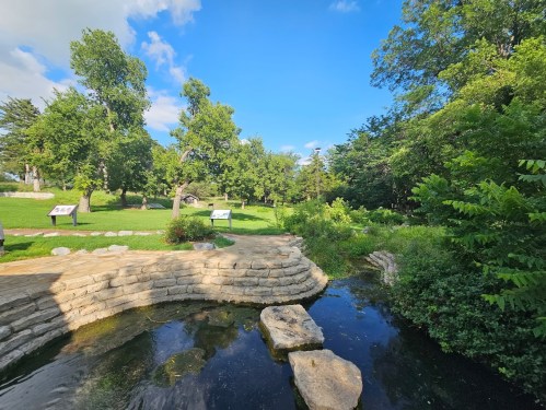 A serene park scene featuring a stone path, a small stream, and lush greenery under a clear blue sky.