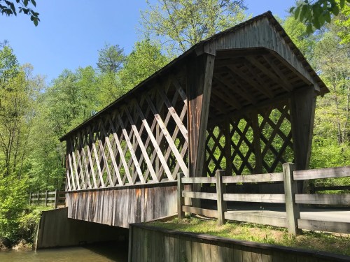 A wooden covered bridge with a lattice design, surrounded by lush green trees and a clear blue sky.