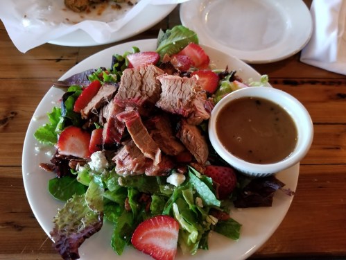 A colorful salad topped with sliced beef, strawberries, and a side of dressing on a wooden table.