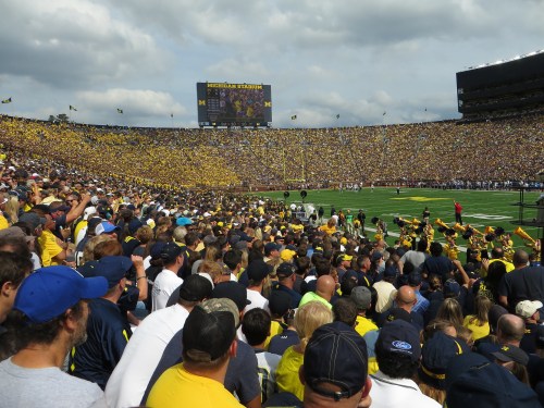 A large crowd in yellow and blue fills Michigan Stadium, with flags waving and a scoreboard visible in the background.