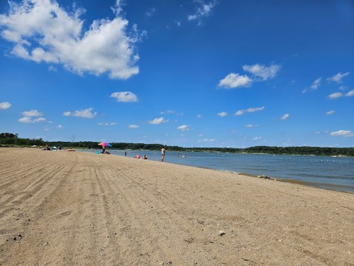A sandy beach with people enjoying the sun, colorful umbrellas, and a calm blue lake under a bright sky with fluffy clouds.