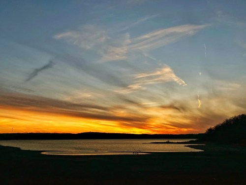 A vibrant sunset over a calm lake, with colorful clouds reflecting in the water and silhouettes of trees in the background.