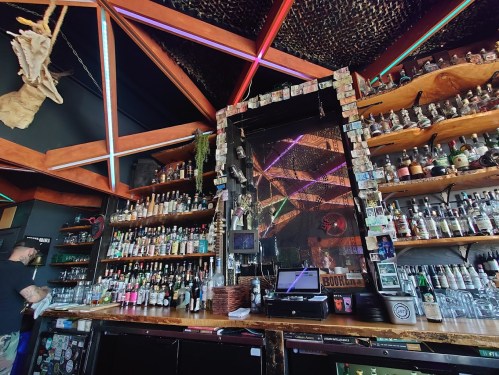 A vibrant bar interior with colorful lighting, shelves filled with bottles, and a bartender preparing drinks.