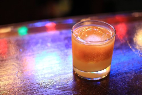 A glass of orange cocktail with a large ice cube, sitting on a bar with colorful lights in the background.
