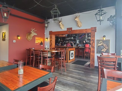 Interior of a rustic restaurant featuring wooden tables, deer head mounts, and a bar in the background.
