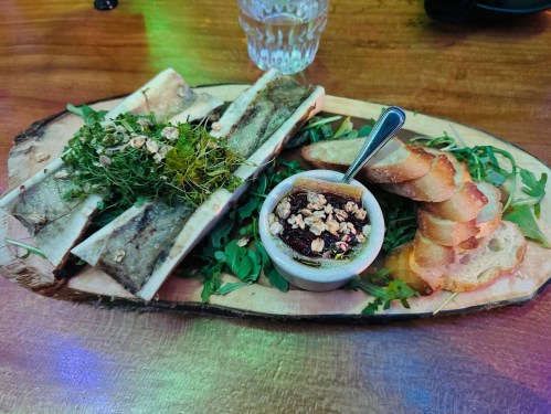 A wooden platter with bone marrow, toasted bread, greens, and a small dish of jam or spread.