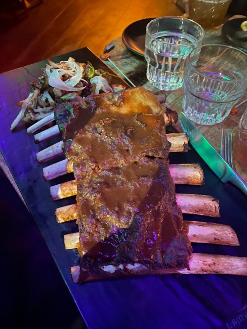 A large rack of ribs with sauce, served on a black platter alongside grilled onions and two glasses of water.