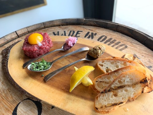 A wooden platter with raw beef tartare, egg yolk, herbs, mustard, lemon, and slices of toasted bread.