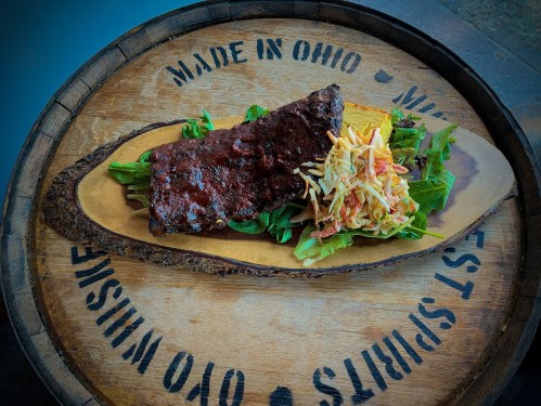 A serving of ribs on a wooden platter, accompanied by greens and coleslaw, with "Made in Ohio" engraved on the edge.