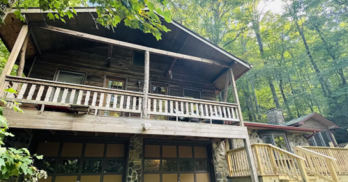 A rustic wooden cabin with a balcony, surrounded by lush green trees and a stone garage below.