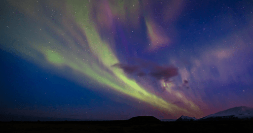 Vibrant auroras dance across a starry night sky, with mountains silhouetted in the foreground.