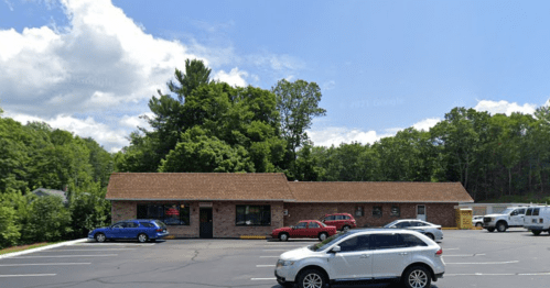 A one-story brick building with a brown roof, surrounded by trees and parked cars in a sunny parking lot.