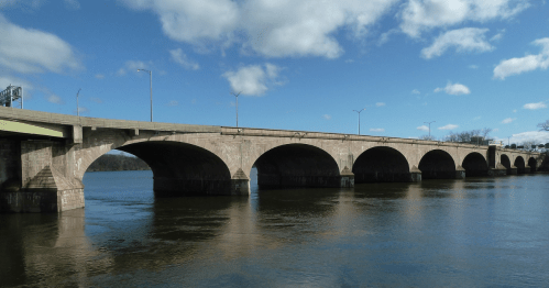 A stone bridge with multiple arches spans a river under a blue sky with scattered clouds.