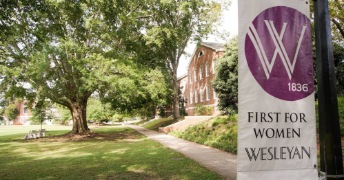 A banner reading "First for Women" hangs near a tree and pathway at Wesleyan College, showcasing the campus's greenery.
