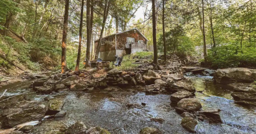 A rustic cabin nestled among trees by a flowing stream, surrounded by rocks and greenery.