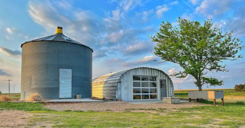 A rustic metal silo next to a modern barn-like structure, set against a blue sky with scattered clouds and green grass.