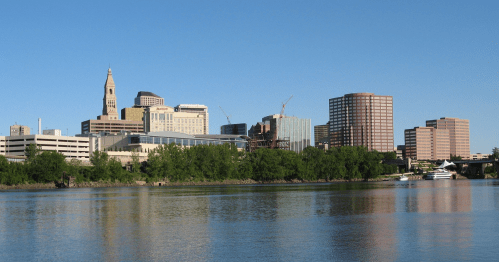 Skyline of Hartford, Connecticut, featuring modern buildings and a clear blue sky reflected in the river.