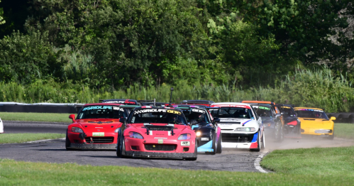 A group of race cars competes on a winding track surrounded by greenery on a sunny day.
