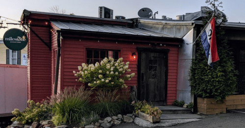 A cozy red building with a wooden door, surrounded by greenery and flags, featuring a sign that reads "milestone."