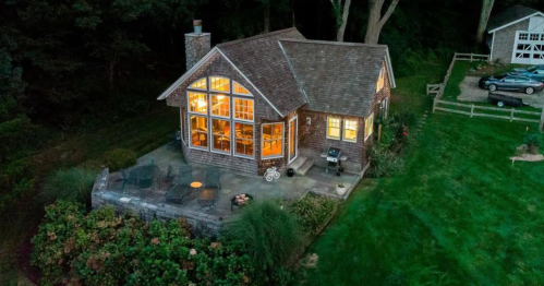 A cozy cottage with large windows, surrounded by greenery and a stone patio, illuminated at dusk.