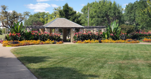 A landscaped garden with colorful flowers and a stone gazebo surrounded by lush greenery under a clear blue sky.