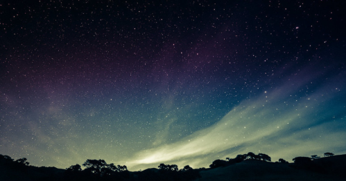 A starry night sky with colorful hues and wispy clouds over a dark landscape.
