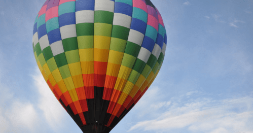 A colorful hot air balloon with a checkered pattern floats against a blue sky with wispy clouds.