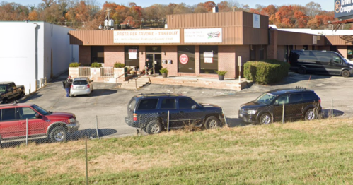 A restaurant with a takeout sign, surrounded by parked cars and greenery, set against a backdrop of autumn trees.