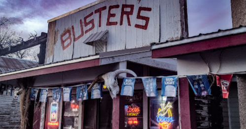 A rustic building with the sign "Busters," featuring neon beer signs and decorative banners.