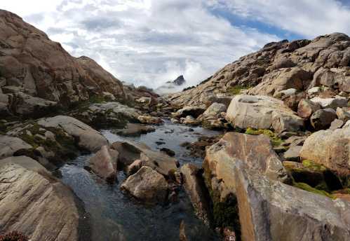 A rocky landscape with a stream flowing through, surrounded by mountains and clouds under a partly cloudy sky.