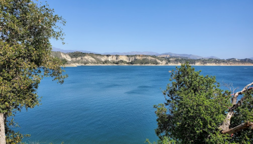 A serene lake surrounded by green trees and distant hills under a clear blue sky.