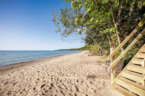 A sandy beach with gentle waves, framed by trees and a wooden staircase leading down to the shore. Clear blue sky above.