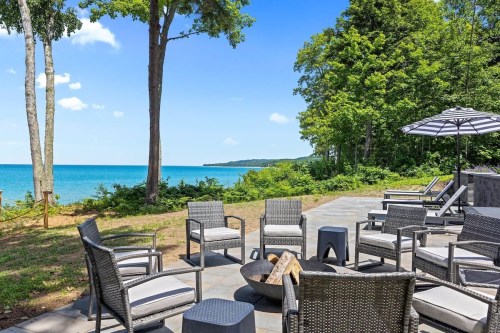 A serene lakeside patio with wicker chairs, a fire pit, and an umbrella, surrounded by trees and clear blue water.