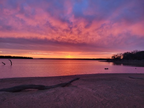 A serene lakeside view at sunset, with vibrant pink and purple clouds reflecting on the water's surface.