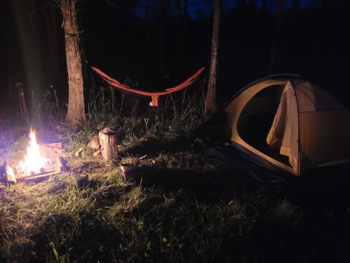 A cozy campsite at night featuring a tent, a hammock, and a crackling campfire surrounded by trees.