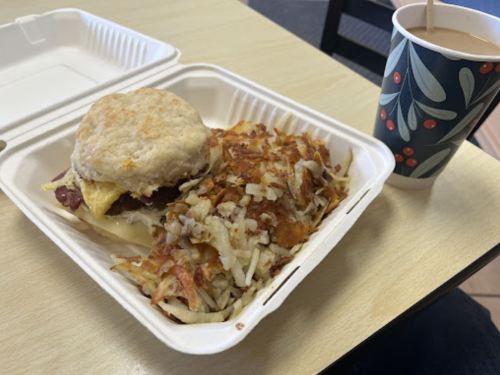 A biscuit sandwich with cheese and meat, served with crispy hash browns and a cup of coffee on a table.
