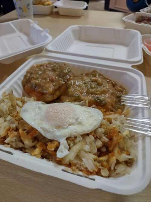 A takeout container with two biscuits covered in gravy, hash browns, and a fried egg on top.