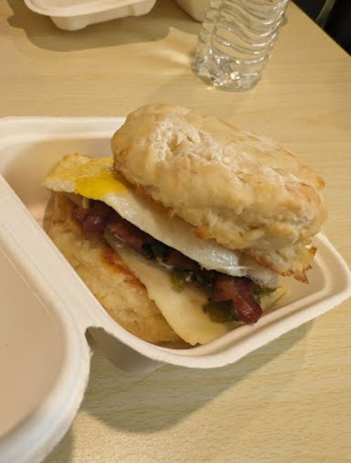 A biscuit sandwich with egg, cheese, and meat, served in a takeout container on a wooden table.