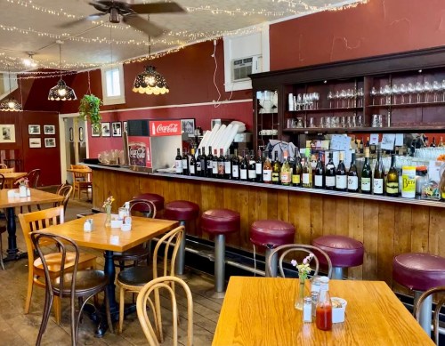Cozy restaurant interior with wooden tables, red leather stools, and a well-stocked bar against a warm, decorated wall.