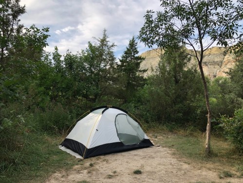 A tent set up in a grassy area surrounded by trees and shrubs, with a rocky hillside in the background.