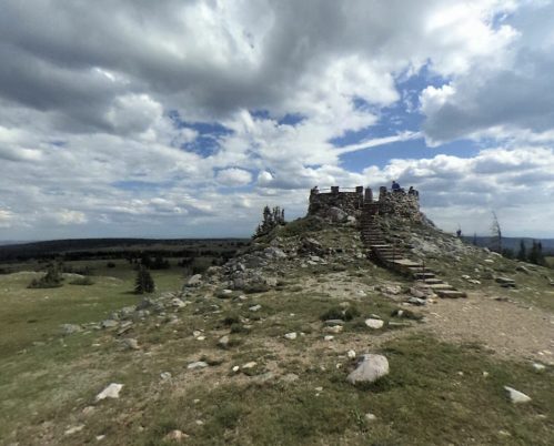 A rocky hill with a stone structure on top, surrounded by grassy fields and a cloudy sky.