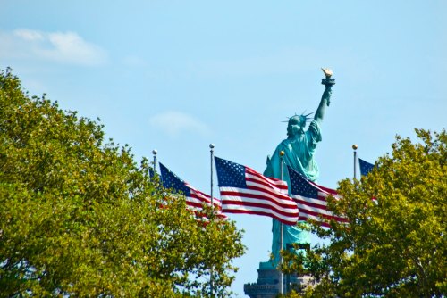 The Statue of Liberty stands tall behind waving American flags and green trees under a clear blue sky.