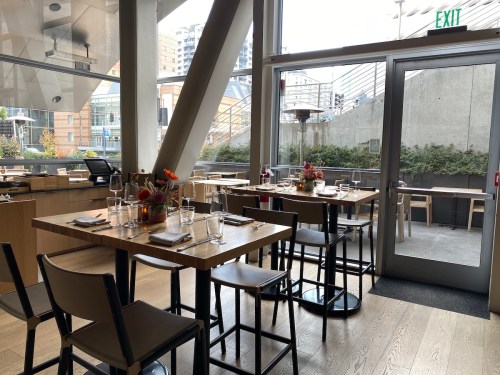 A modern restaurant interior with wooden tables, chairs, and large windows, featuring floral centerpieces and outdoor access.