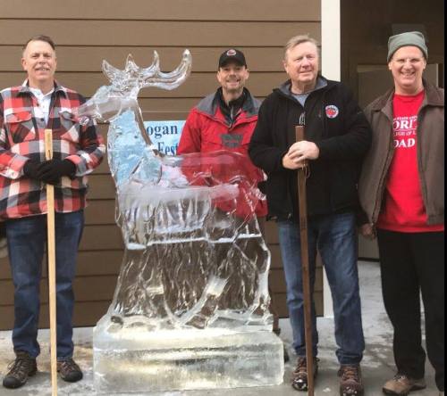 Four men stand beside a large ice sculpture of a moose, holding walking sticks, in a festive outdoor setting.